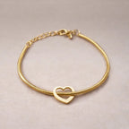 Zoom sur le bracelet "Coeur" en acier doré, orné d'un cœur mobile, posé sur un fond beige texturé.