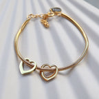 Zoom sur le bracelet "Emotive" en acier doré, orné de deux cœurs mobiles, posé sur un fond beige texturé.