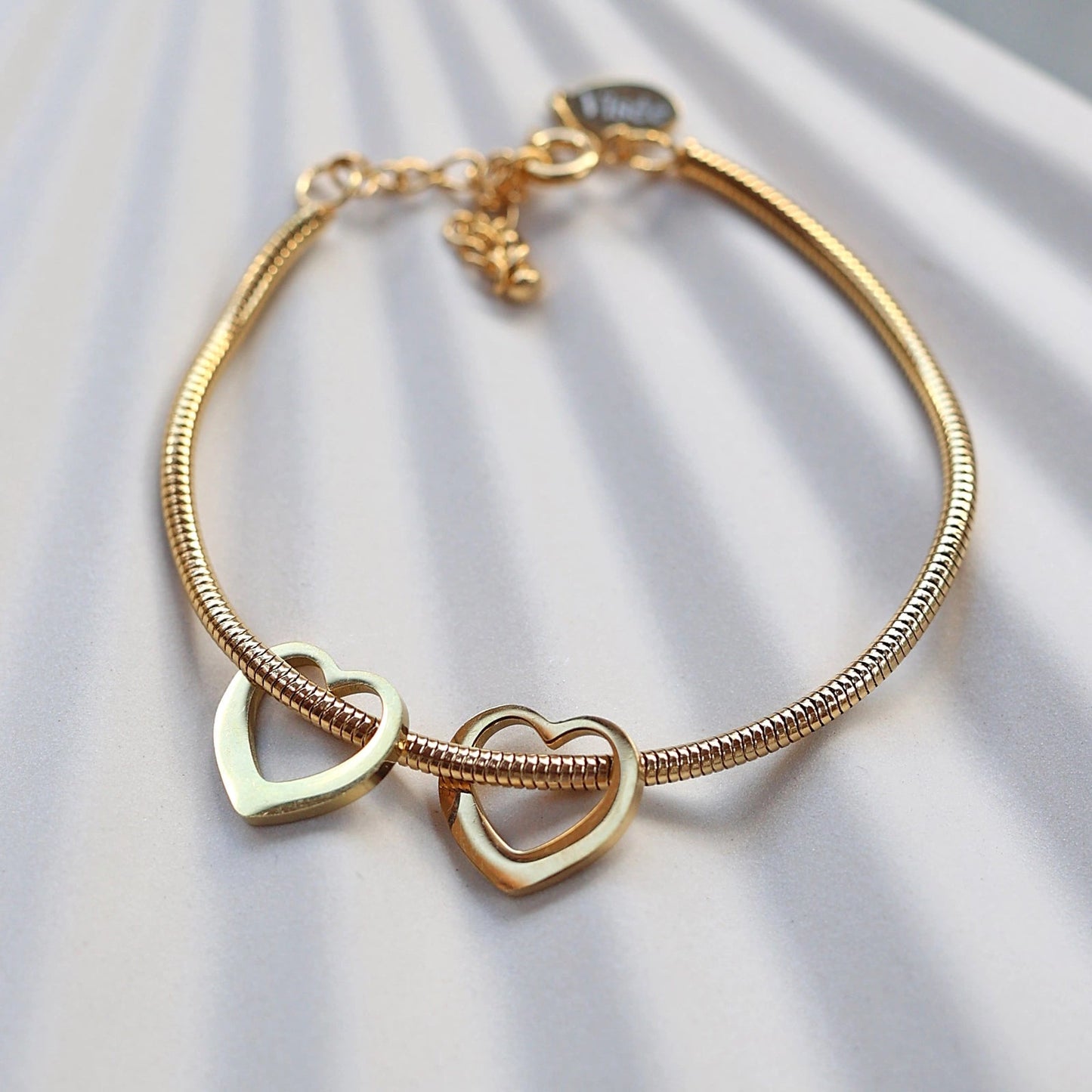 Zoom sur le bracelet "Emotive" en acier doré, orné de deux cœurs mobiles, posé sur un fond beige texturé.