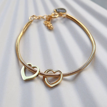 Zoom sur le bracelet "Emotive" en acier doré, orné de deux cœurs mobiles, posé sur un fond beige texturé.