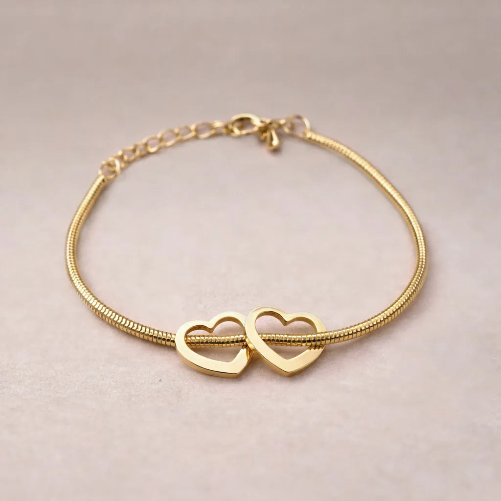 Zoom sur le bracelet "Coeurs" en acier doré, orné de deux coeurs mobiles, posé sur un fond beige texturé.