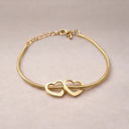 Zoom sur le bracelet "Coeurs" en acier doré, orné de deux coeurs mobiles, posé sur un fond beige texturé.