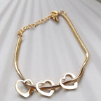 Zoom sur le bracelet "Emotive" en acier doré, orné de trois cœurs mobiles, posé sur un fond beige texturé.