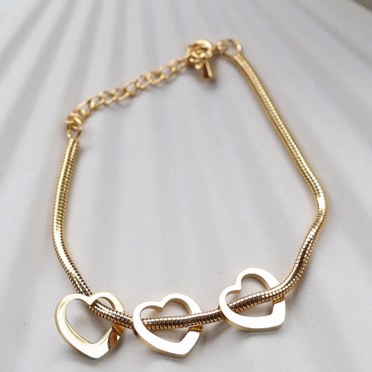 Zoom sur le bracelet "Emotive" en acier doré, orné de trois cœurs mobiles, posé sur un fond beige texturé.