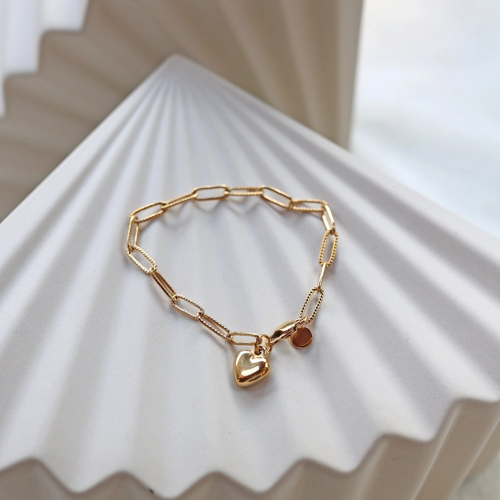 Bracelet Valeur acier inoxydable doré, avec breloque cœur personnalisable, posé sur un fond beige texturé.