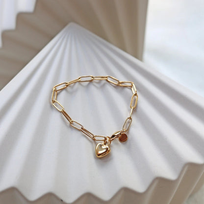 Bracelet Valeur acier inoxydable doré, avec breloque cœur personnalisable, posé sur un fond beige texturé.