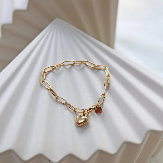 Bracelet Valeur acier inoxydable doré, avec breloque cœur personnalisable, posé sur un fond beige texturé.