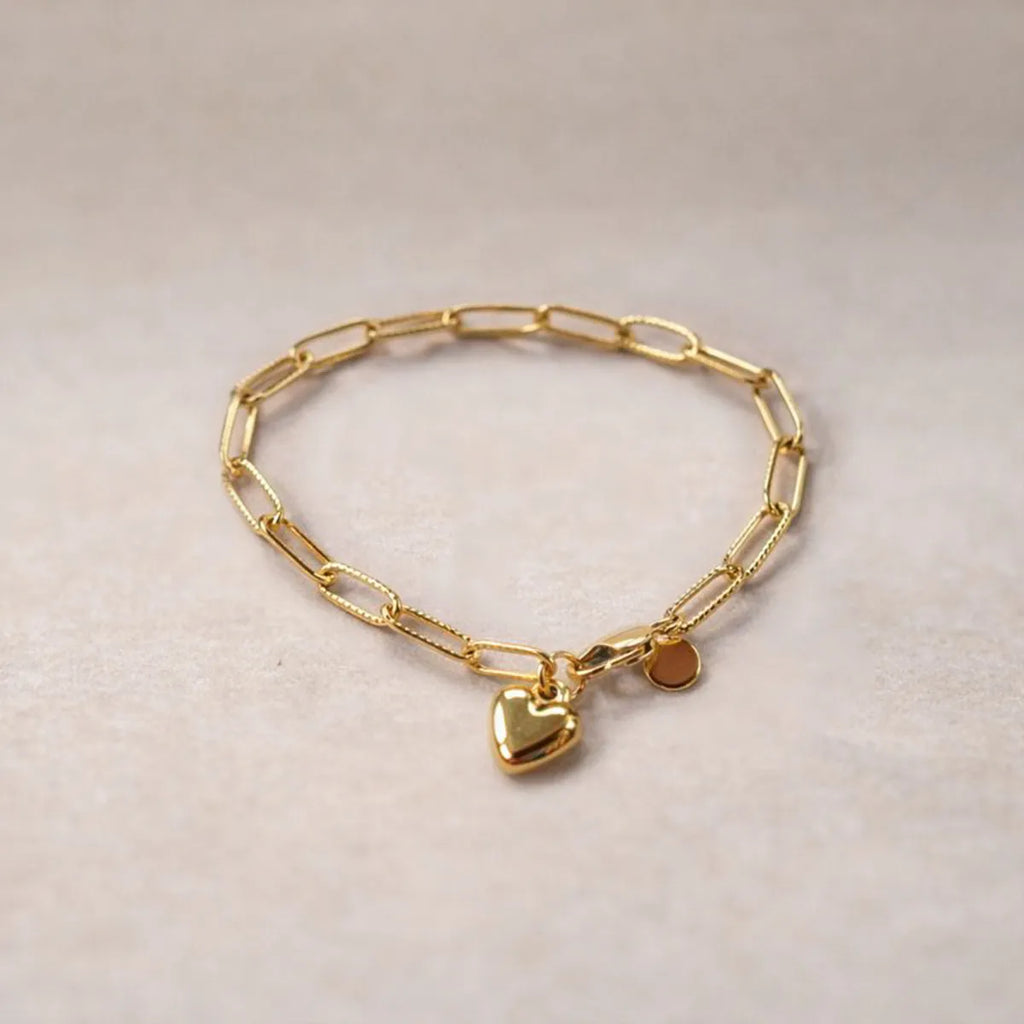 Bracelet Valeur acier inoxydable doré, avec breloque cœur personnalisable, posé sur un fond beige texturé.