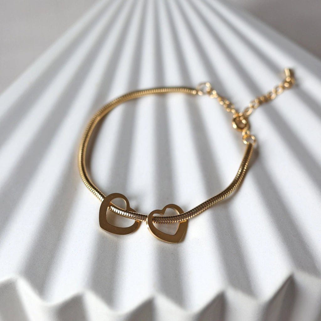 Bracelet "Emotive" en acier doré, orné de trois cœurs mobiles, posé sur un fond beige texturé.