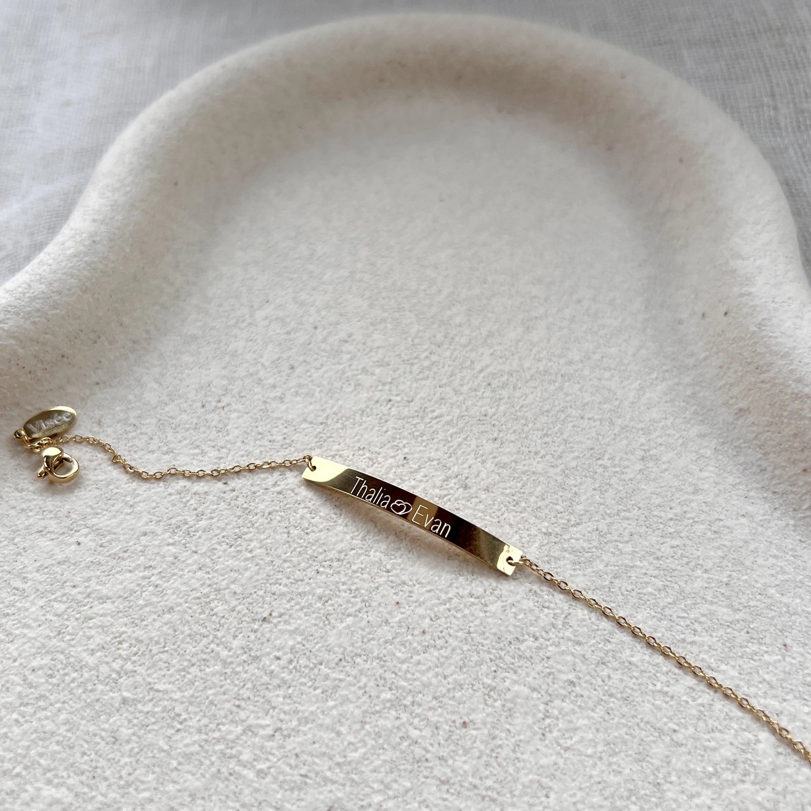 Bracelet gourmette Lien en acier inoxydable doré,  personnalisé de noms "Thalia Evan", posé sur un fond beige texturé.
