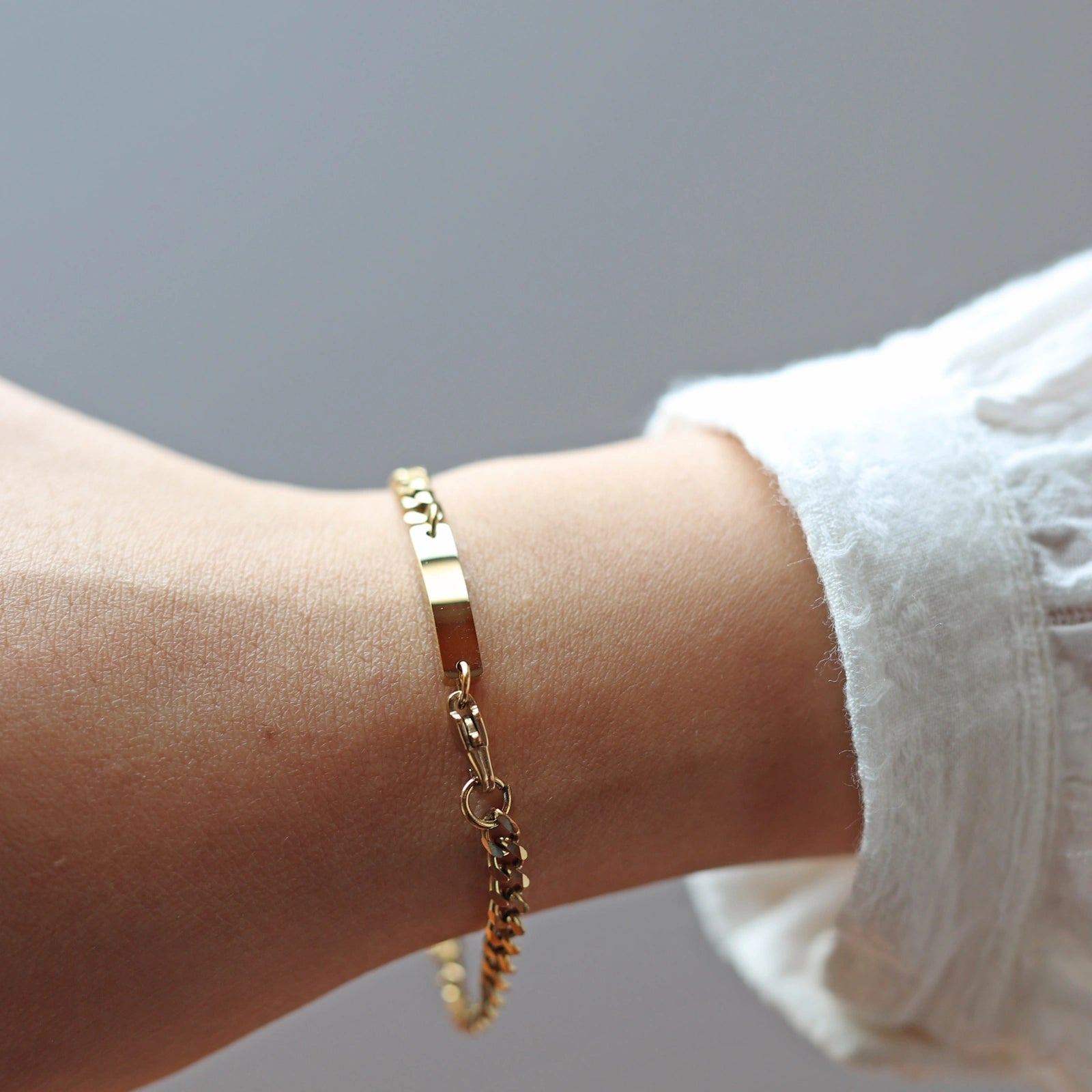 Bracelet Force en acier doré, style gourmette, avec plaque gravable, porté autour du poignet. Le mannequin porte une chemise blanche ajourée.