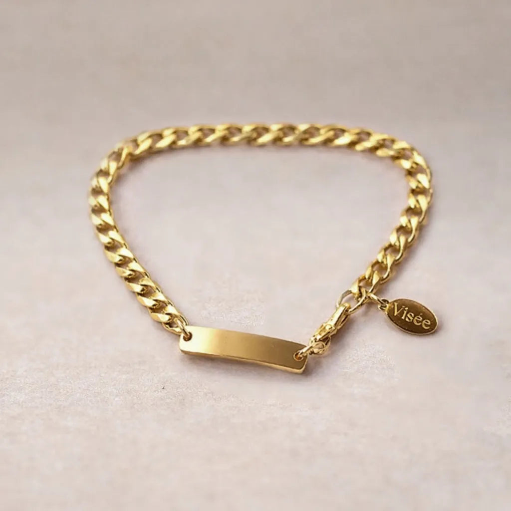 Bracelet "Libère toi" en acier doré, style gourmette, avec plaque gravable, posé sur un fond beige texturé.