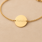 Zoom sur le bracelet "Aime", médaille ronde gravée au nom de Nathalie,en acier doré, photographié sur fond beige texturé.