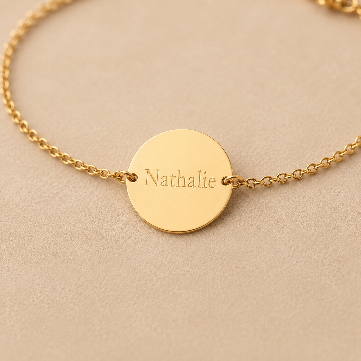 Zoom sur le bracelet "Aime", médaille ronde gravée au nom de Nathalie,en acier doré, photographié sur fond beige texturé.