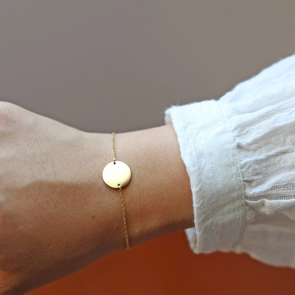Bracelet Aimé en acier inoxydable doré, personnalisable avec médaille ronde et chaine fine, porté sur le poignet de notre mannequin, habillé d'une chemise blanche avec broderie. 