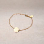 Zoom sur le bracelet Aimé en acier doré personnalisable avec médaille ronde et une chaine fine,  présenté sur un tissu beige texturé. 