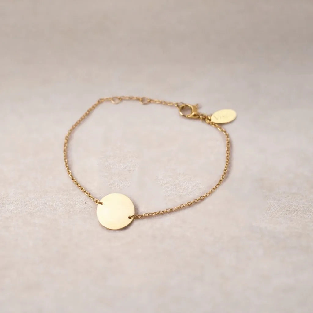 Zoom sur le bracelet "Confiance" en acier doré personnalisable avec médaille ronde et une chaine fine,  présenté sur un tissu beige texturé. 