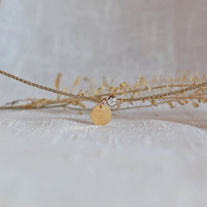 Bracelet "Mon choix" avec médaillon doré personnalisable et pierre blanche, posé sur un fond beige texturé.
