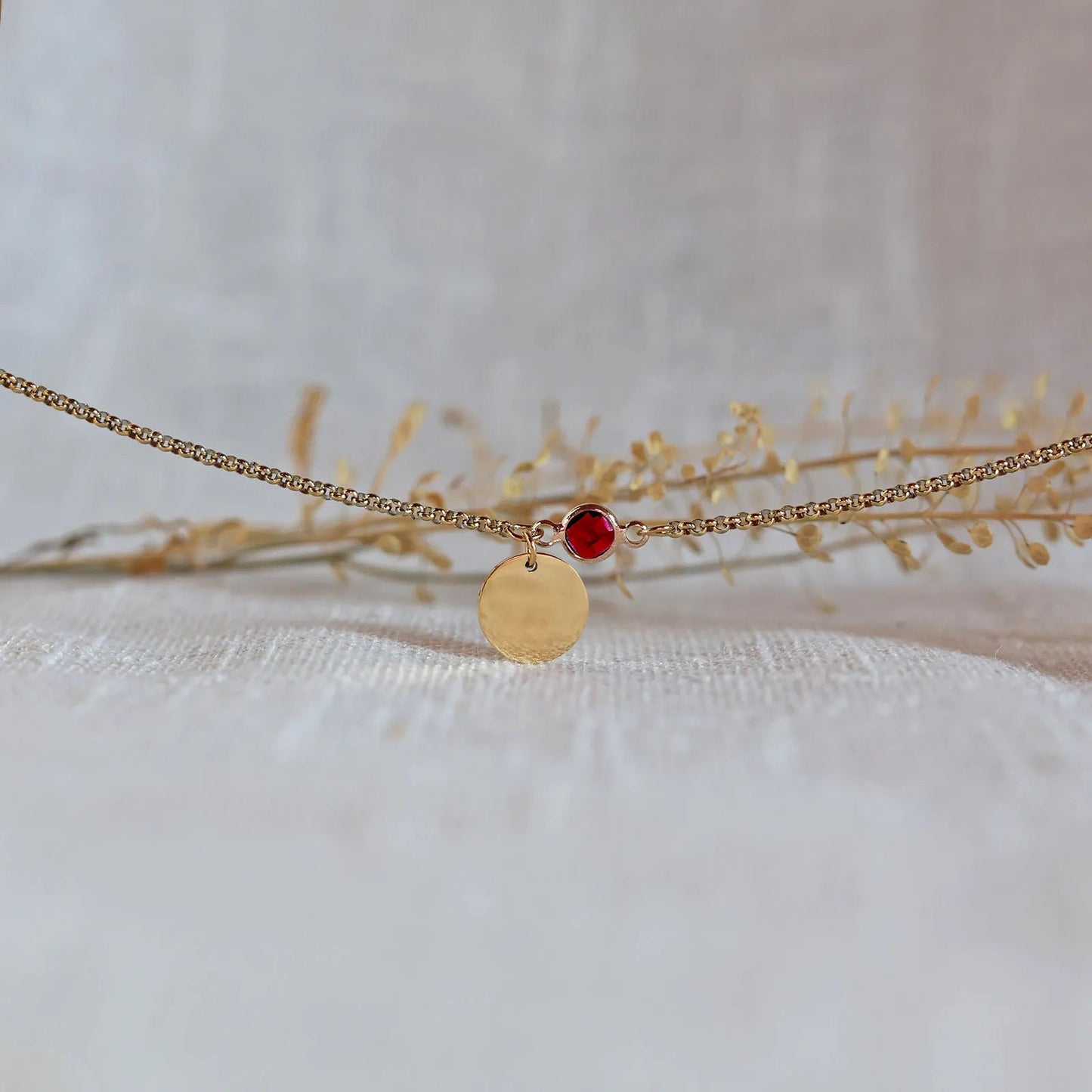 Bracelet "Mon choix" avec médaillon doré personnalisable et pierre rouge, posé sur un fond beige texturé.