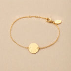 Zoom sur le bracelet "Aime" en acier doré personnalisable avec médaille ronde et une chaine fine , présenté sur un tissu beige texturé. 