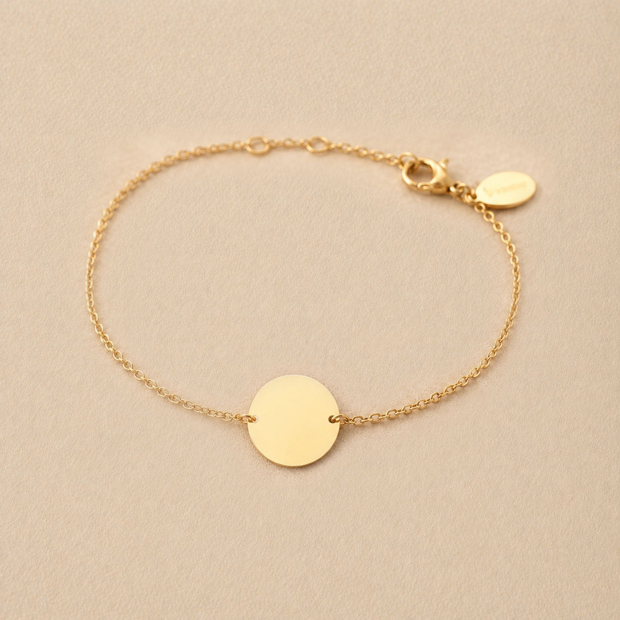 Zoom sur le bracelet "Aime" en acier doré personnalisable avec médaille ronde et une chaine fine , présenté sur un tissu beige texturé. 