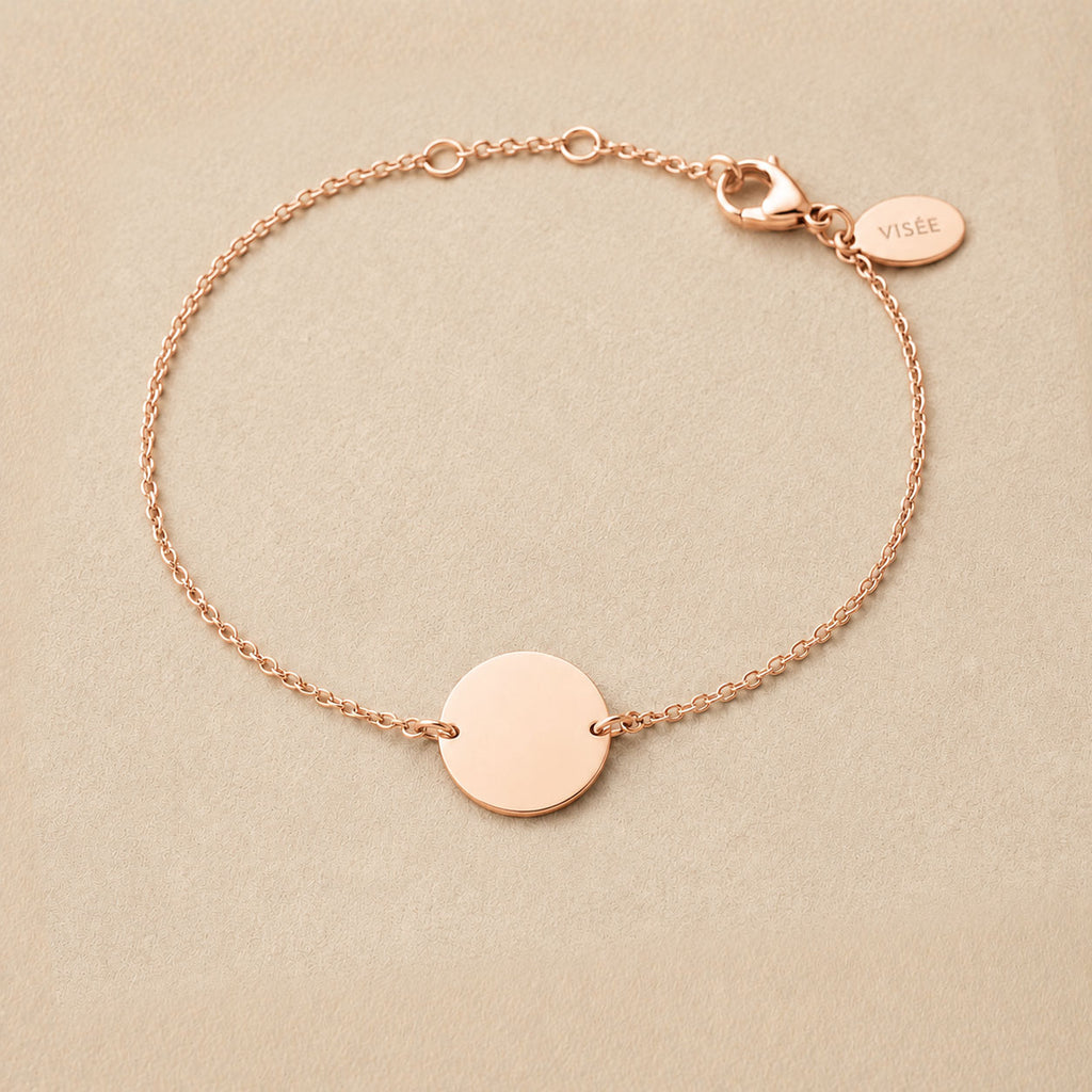 Zoom sur le bracelet "Aime" en acier or rose personnalisable avec médaille ronde et une chaine fine , présenté sur un tissu beige texturé. 