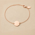 Zoom sur le bracelet "Aime" en acier or rose personnalisable avec médaille ronde et une chaine fine , présenté sur un tissu beige texturé. 