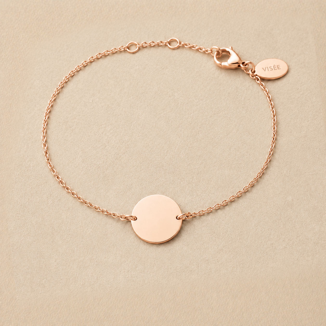 Zoom sur le bracelet "Aime" en acier or rose personnalisable avec médaille ronde et une chaine fine , présenté sur un tissu beige texturé. 
