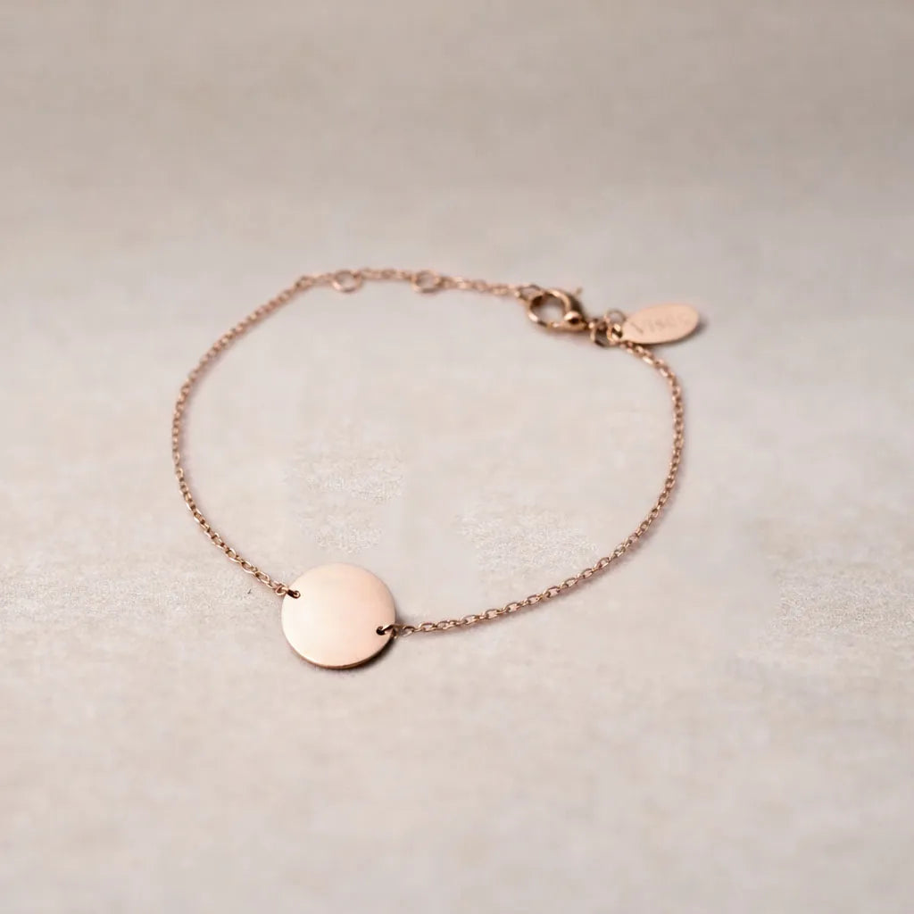 Zoom sur le bracelet "Aime" en acier or rose personnalisable avec médaille ronde et une chaine fine , présenté sur un tissu beige texturé. 