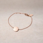 Zoom sur le bracelet "Aime" en acier or rose personnalisable avec médaille ronde et une chaine fine , présenté sur un tissu beige texturé. 