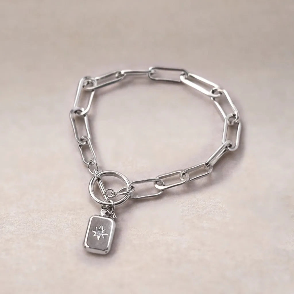 Zoom sur le bracelet "Révélation" en acier argenté avec breloque rectangulaire personnalisable vue de dos, posé sur un fond beige texturé.