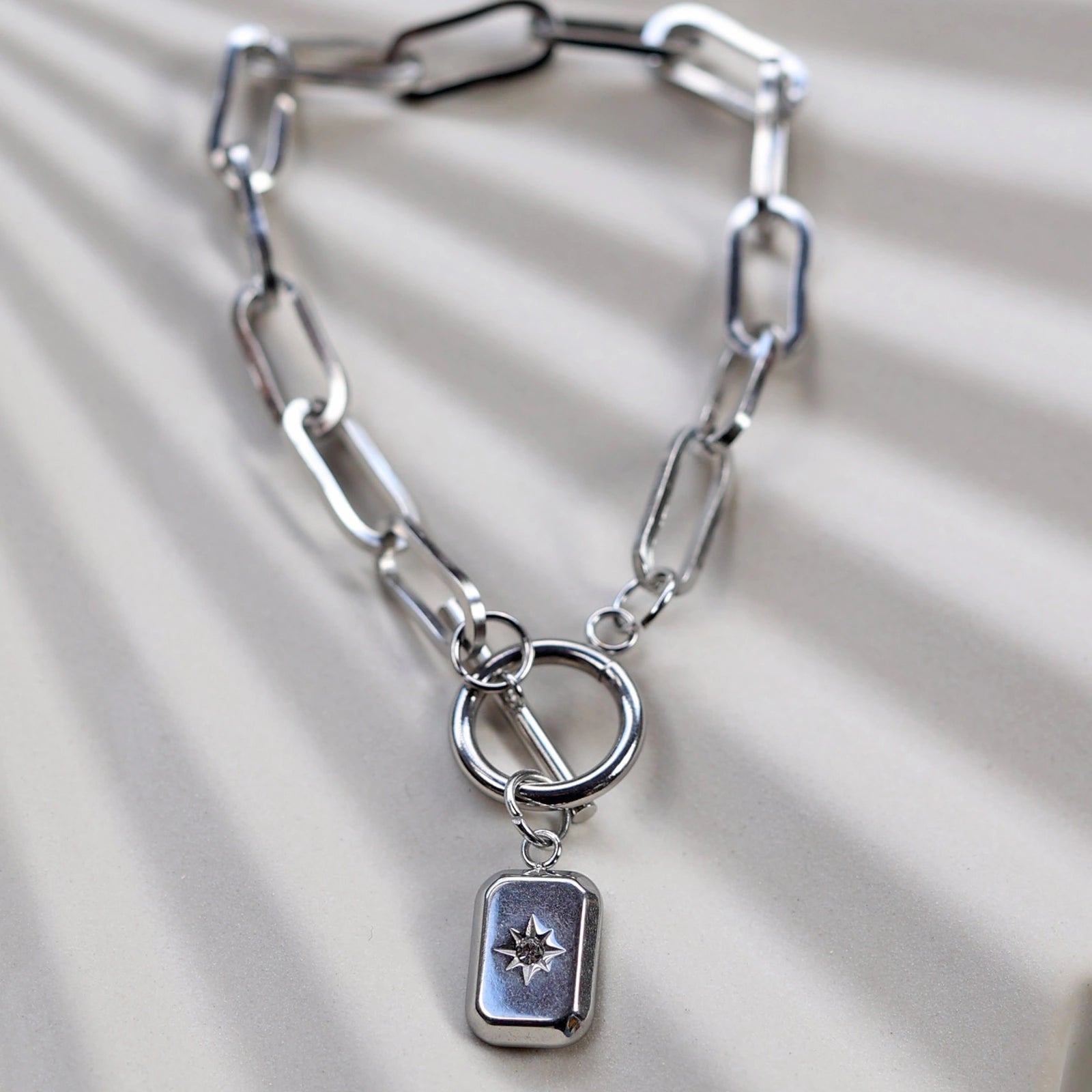 Zoom sur le bracelet "Révélation" en acier argenté, orné d'une breloque rectangulaire personnalisable, posé sur un fond beige texturé.