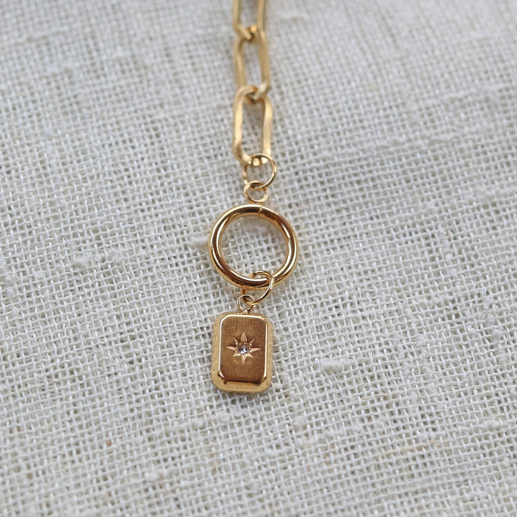 Zoom sur la breloque rectangulaire personnalisable en acier doré, posé sur un fond beige texturé.