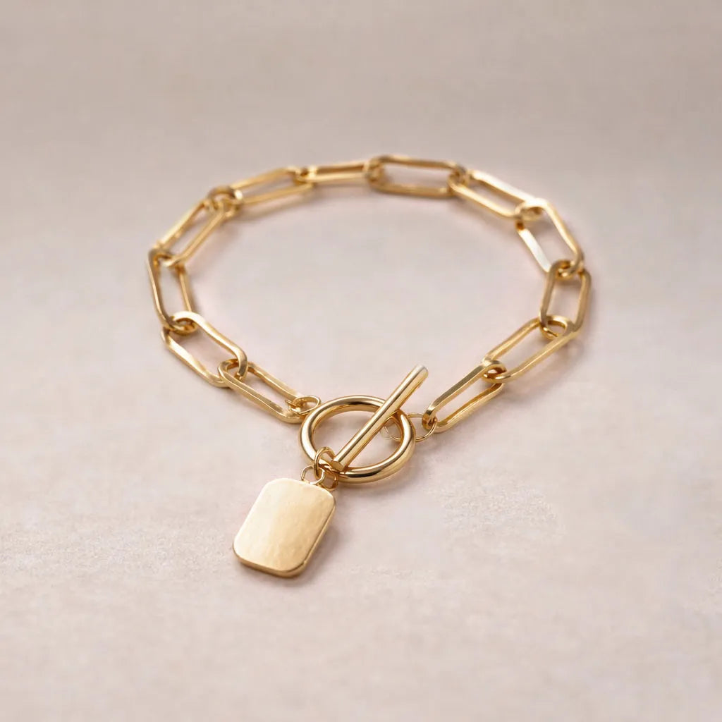 Zoom sur le bracelet "Voeu" en acier doré avec breloque rectangulaire personnalisable vue de dos, posé sur un fond beige texturé.