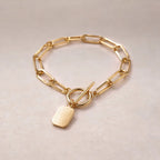 Zoom sur le bracelet "Voeu" en acier doré avec breloque rectangulaire personnalisable vue de dos, posé sur un fond beige texturé.