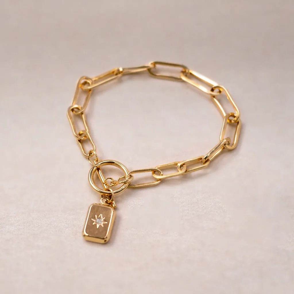 Zoom sur le bracelet Voeu en acier doré avec breloque rectangulaire personnalisable, posé sur un fond beige texturé.