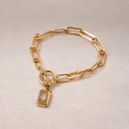 Zoom sur le bracelet Voeu en acier doré avec breloque rectangulaire personnalisable, posé sur un fond beige texturé.