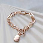 Bracelet "Révélation" en acier rosé avec breloque rectangulaire personnalisable, posé sur un fond beige texturé.