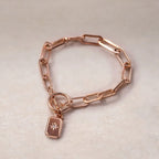 Zoom sur le bracelet Voeu en acier or rose avec breloque rectangulaire personnalisable vue de dos, posé sur un fond beige texturé.