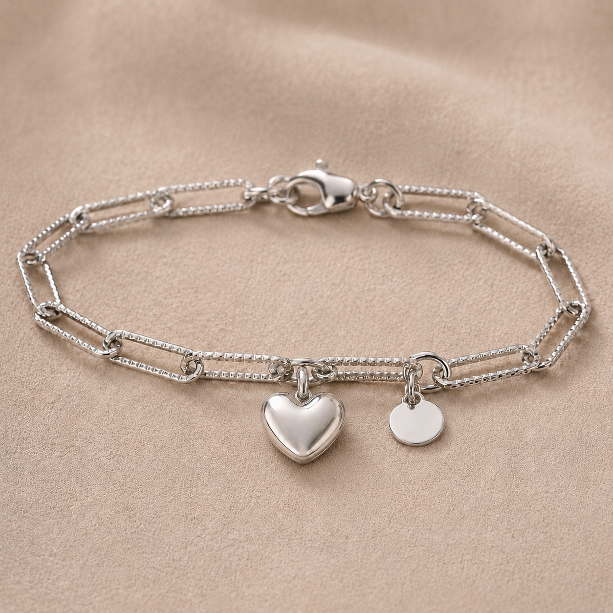 Bracelet Attache personnalisable avec initial en acier inoxydable argenté, avec breloque cœur , posé sur un fond beige texturé, en packshoot