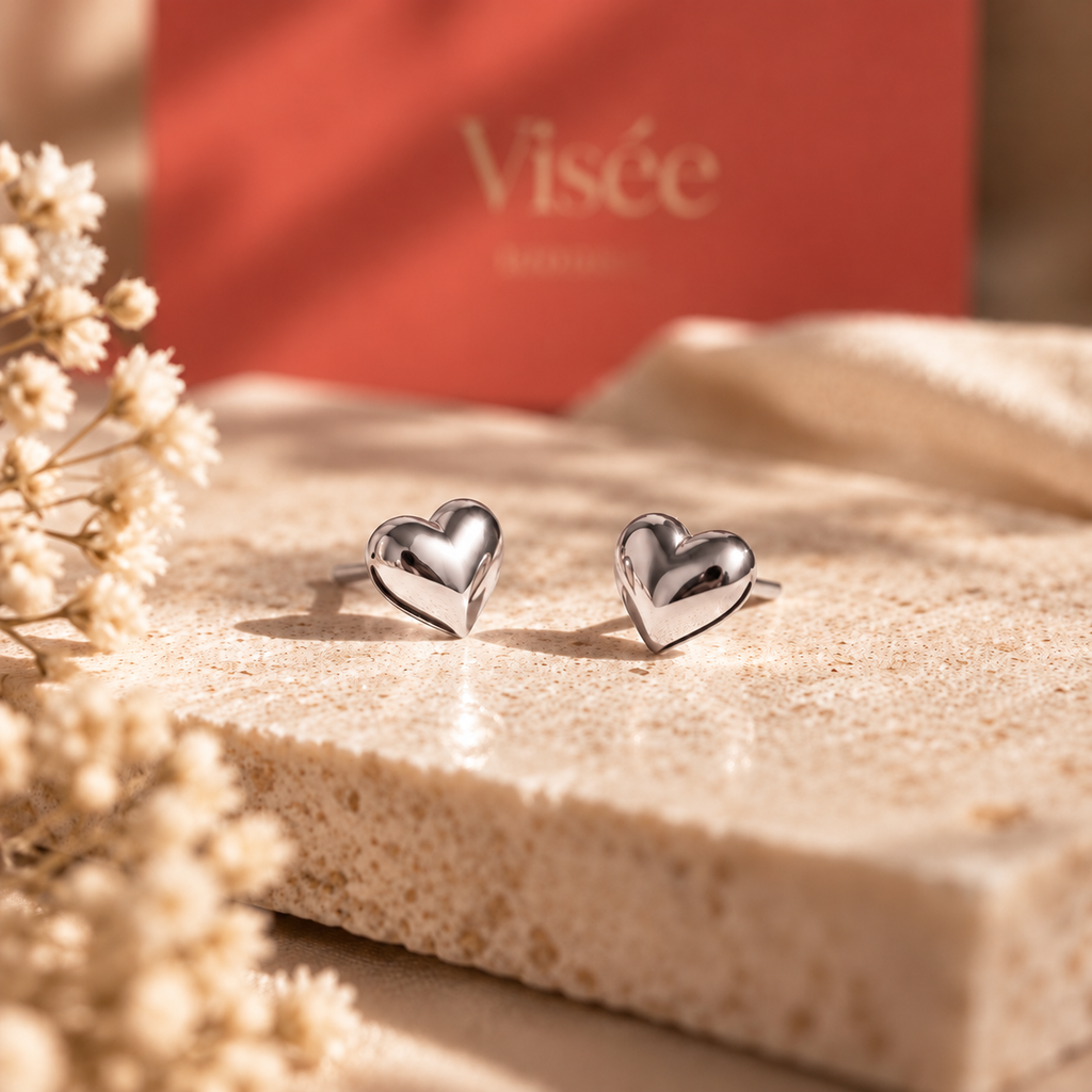 Boucles d'oreilles puces cœur bombé argentées du Coffret Précieux Visée, posées sur une pierre claire devant l'écrin