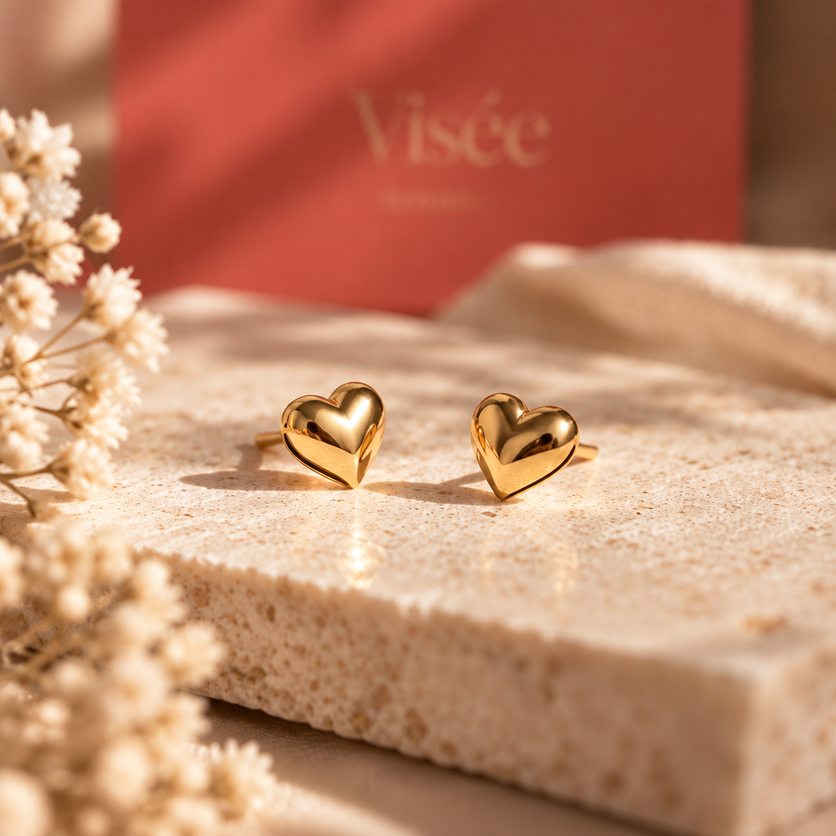 Boucles d'oreilles puces cœur bombé dorées du Coffret Précieux Visée, posées sur une pierre claire devant l'écrin