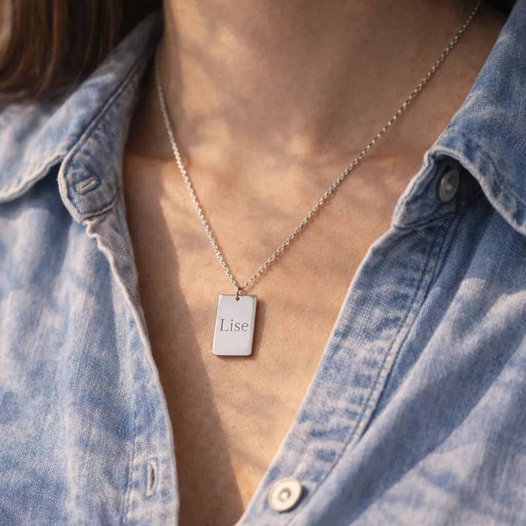 Collier Tracé en acier argenté, avec médaille rectangulaire gravé du prénom de Lise, porté autour du cou. Le modèle porte une chemise en jeans.