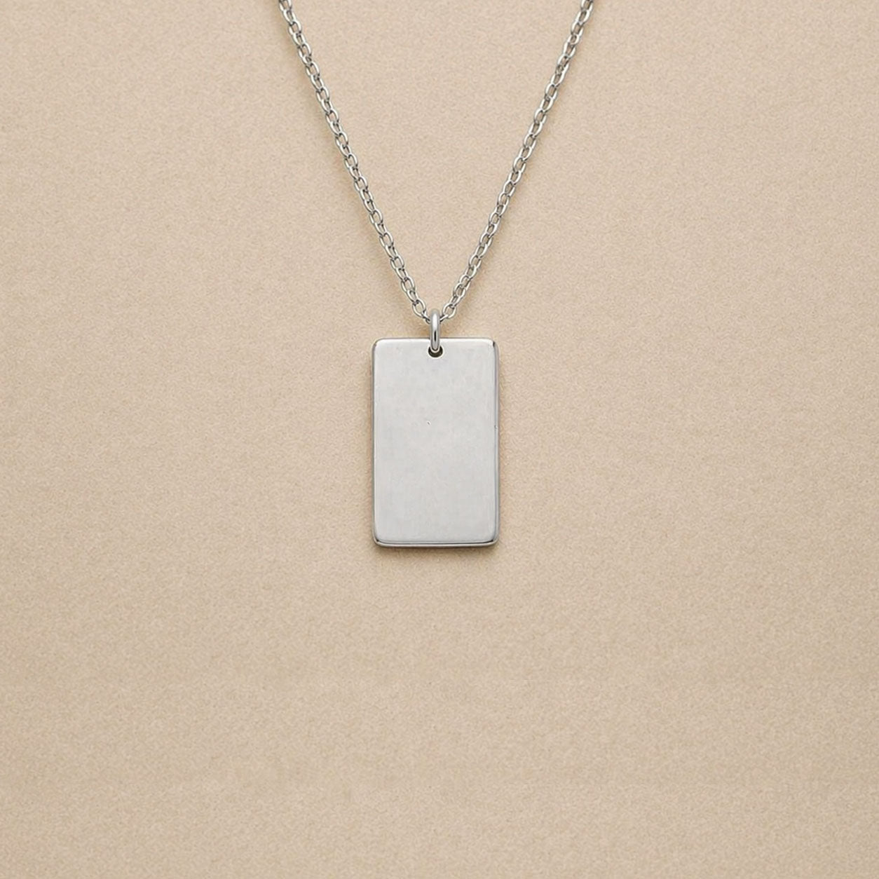 Collier Trace plaque rectangulaire personnalisable en acier inoxydable argenté sur fond beige. 