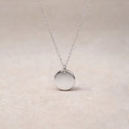 Collier "Reflet" avec médaille ronde personnalisé en acier inoxydable argenté, posé sur un fond beige texturé