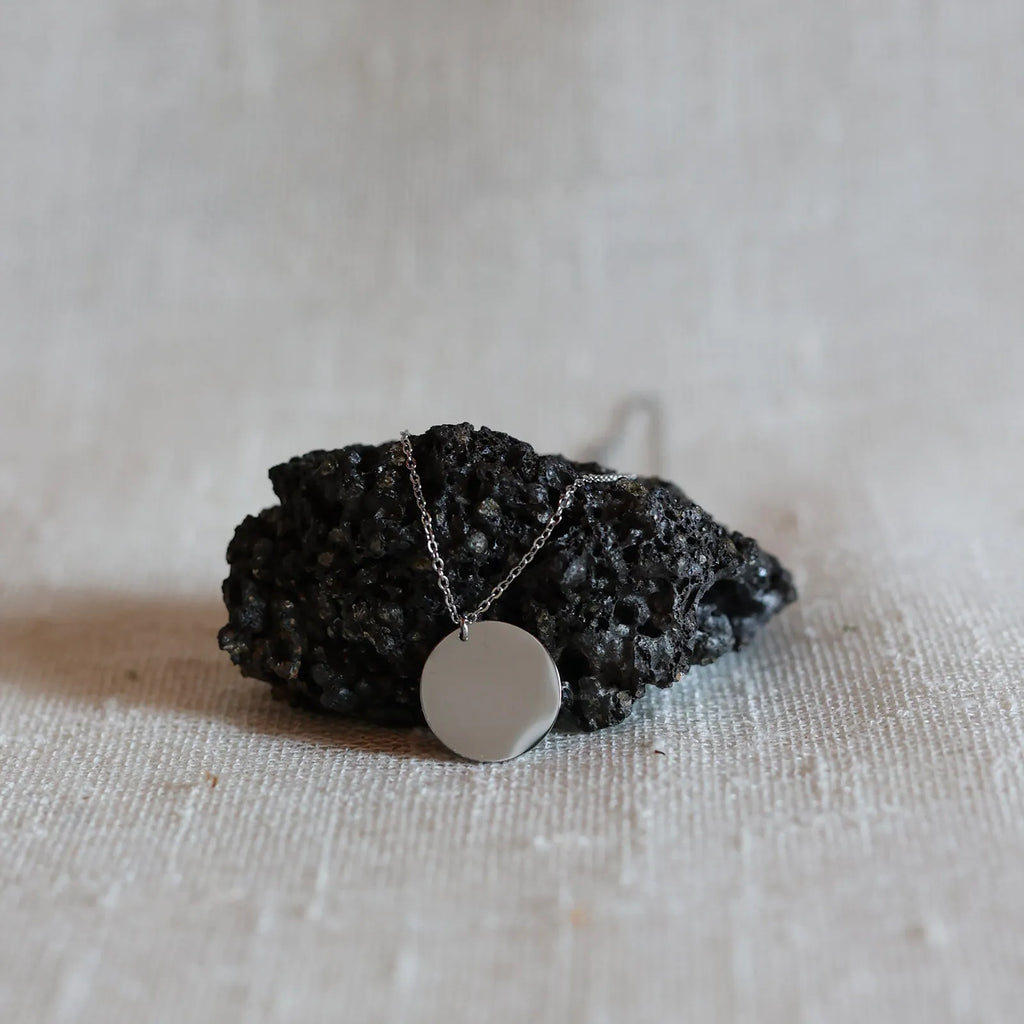 Collier Renaissance argent, médaille ronde martelée à graver de dos, photographié posée sur une pierre volcanique sur un fond beige texturé. 