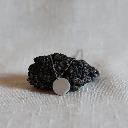 Collier Renaissance argent, médaille ronde martelée à graver de dos, photographié posée sur une pierre volcanique sur un fond beige texturé. 