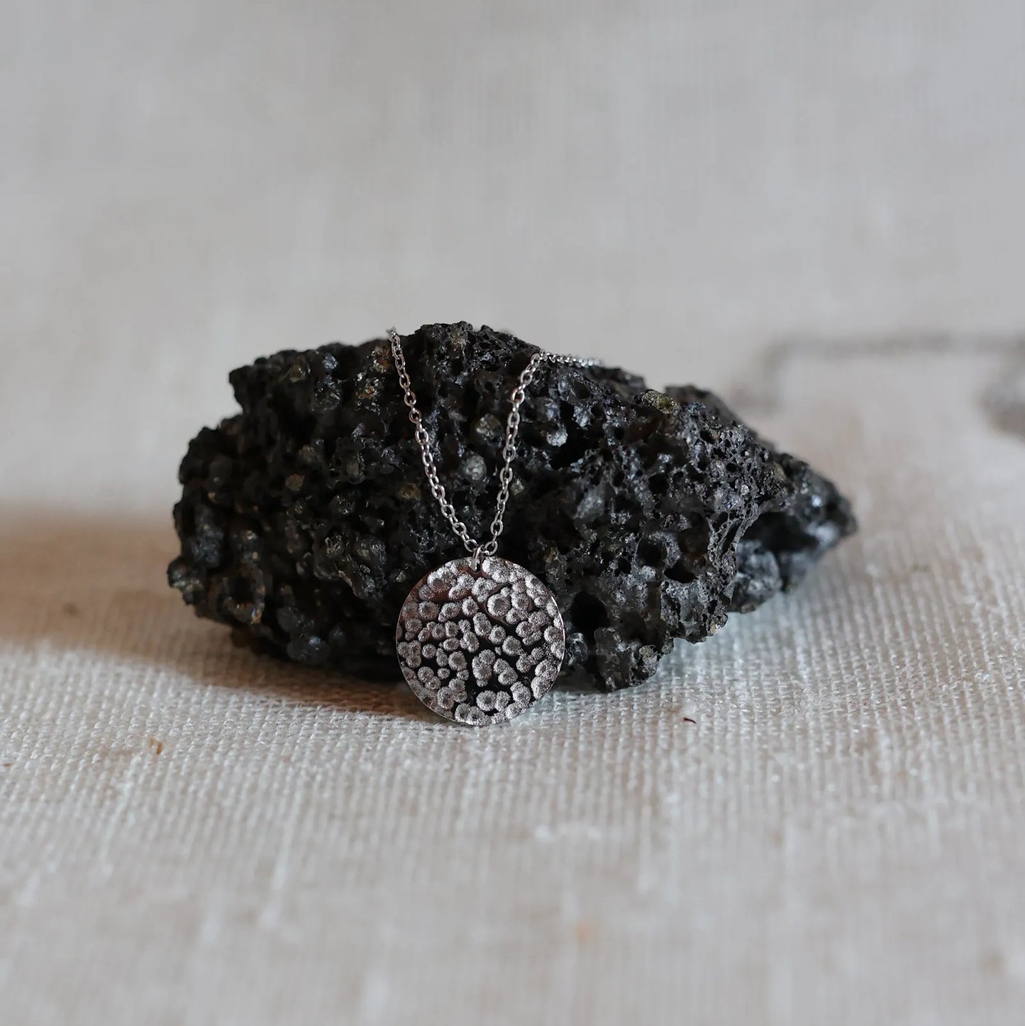 Collier Renaissance argent, médaille ronde martelée à graver, photographié posée sur une pierre volcanique sur un fond beige texturé. 