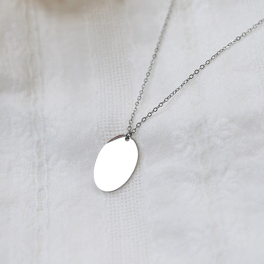 Collier "Tendresse", pendentif ovale personnalisable en acier inoxydable argenté, présenté sur un fond beige texturé.