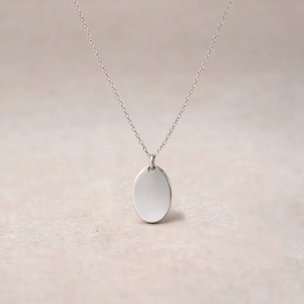 Collier "Tendresse", pendentif ovale personnalisable en acier inoxydable argenté, présenté sur un fond beige texturé.